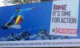 A Philip Morris International advertisement which states, 'Maybe it's time to action. Be Marlboro,' with the word Maybe crossed through.  It shows a totally airbourne snow-boarder holding onto the board with one hand, with the peaks of snow covered mountains in the background.