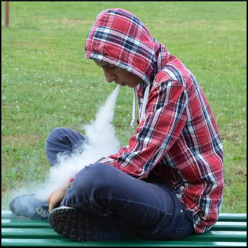 Young hooded man sitting cross legged on a bench while vaping. 